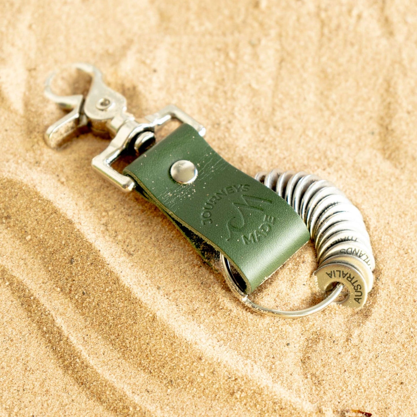 Forest green leather travel keychain with engraved destination tokens on sandy surface.