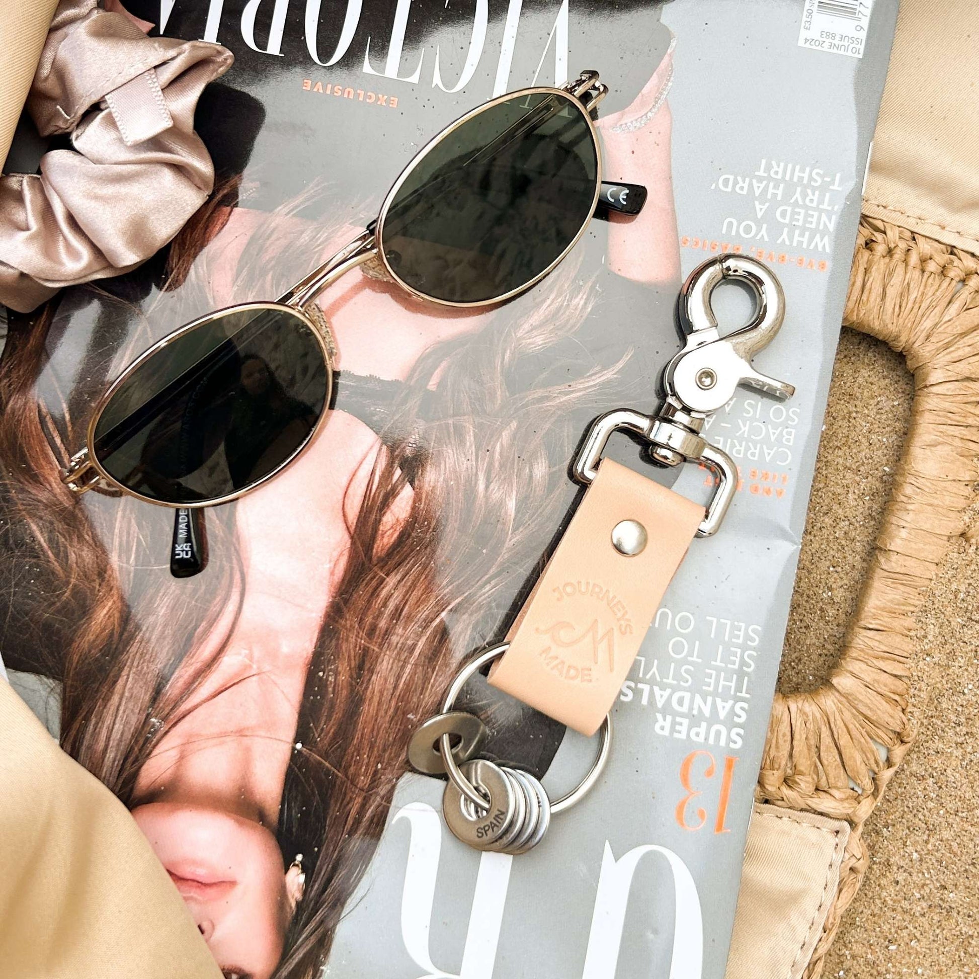 natural travel gift with leather travel keyring and engraved travel tokens laying on a Grazia magazine with a pair of black sunglasses in an open beach bag