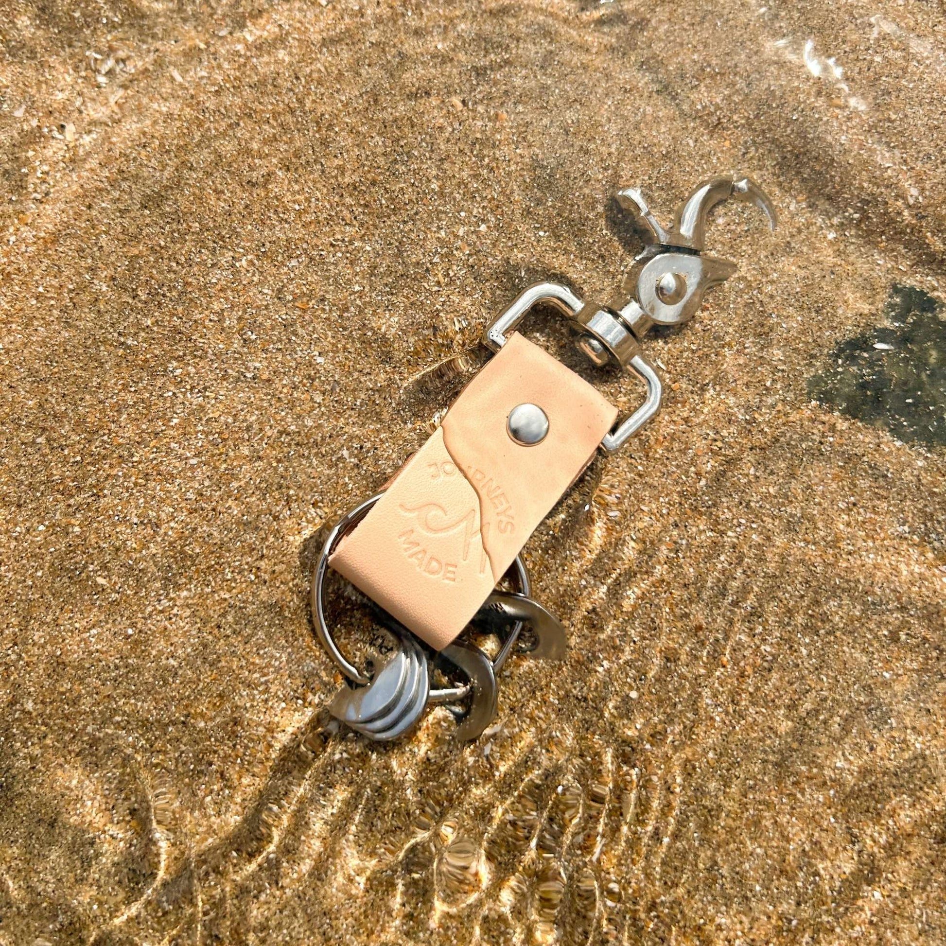 Natural leather travel keychain resting in shallow water on a sandy beach, symbolizing adventure and travel.