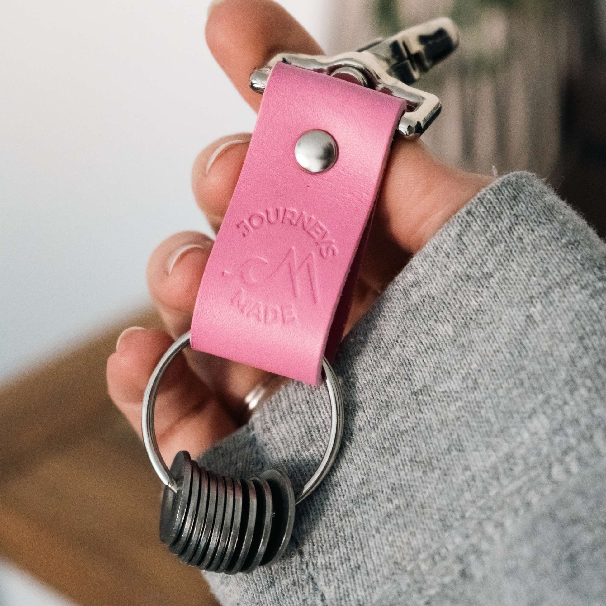 pink travel gift consisting of leather travel keyring and engraved travel tokens on the split ring being held by a white woman's hand wearing a grey hooded sweatshirt
