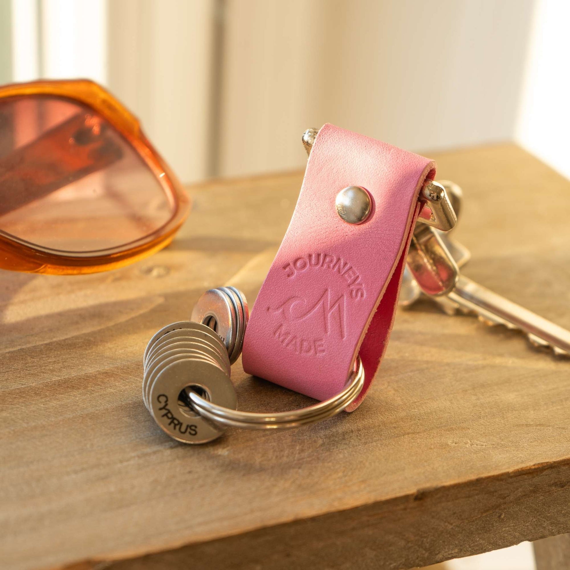 pink travel gift consisting of leather travel keyring and engraved travel tokens on the split ring on a wooden table at sunset with a pair of sunglasses in the background