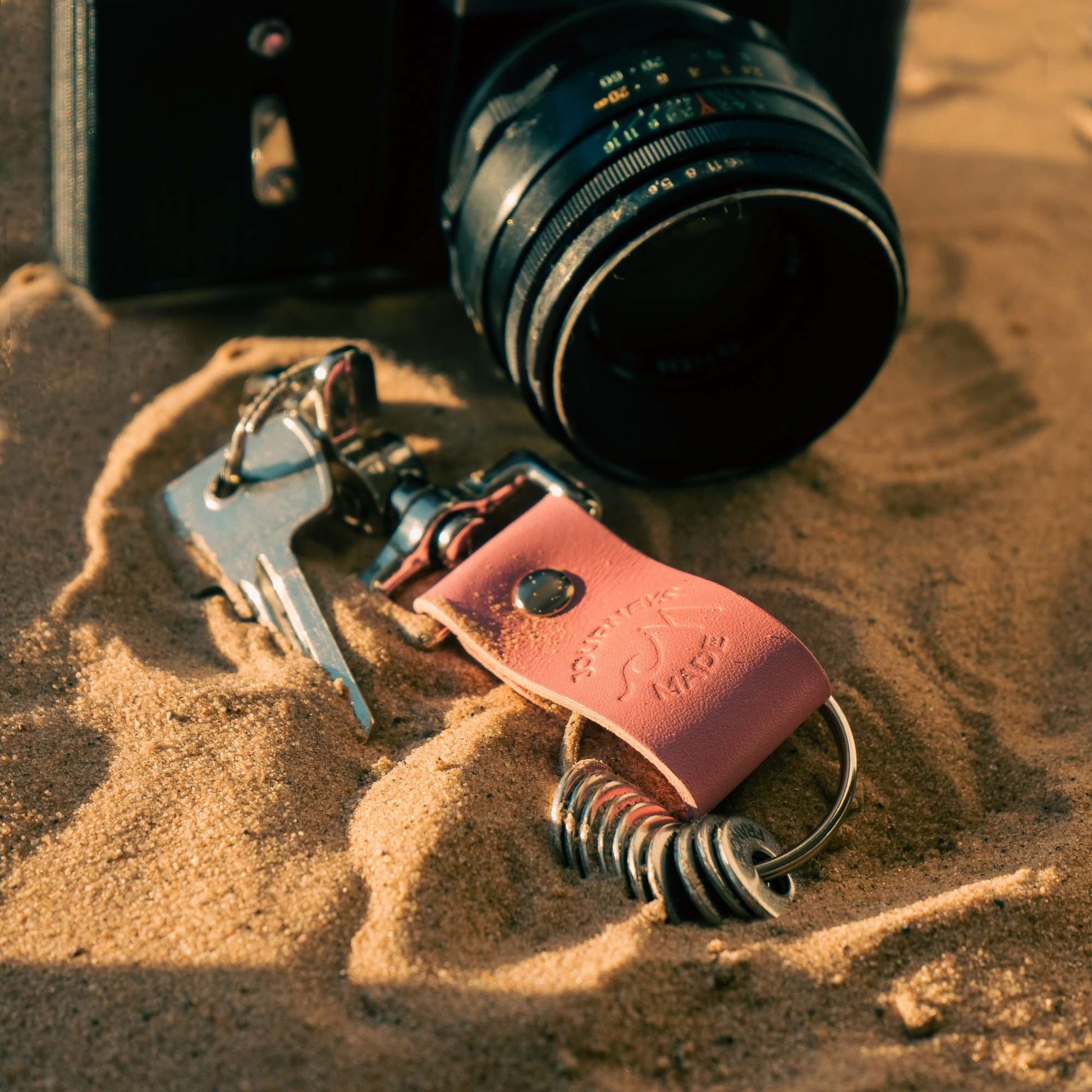 pink travel gift consisting of leather travel keyring and engraved travel tokens on the split ring laying on a bed of sand with a black camera in the background at sunset