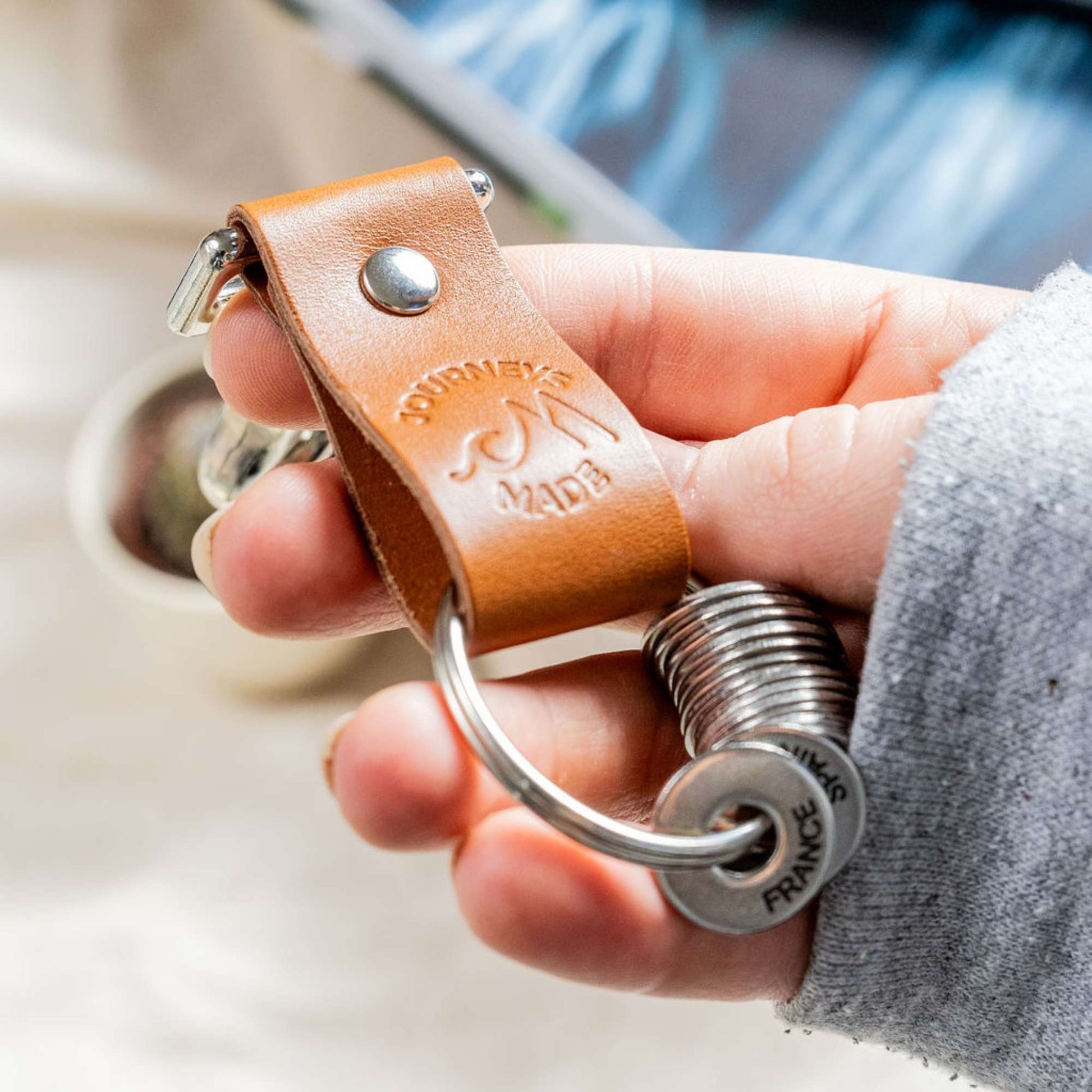 tan travel gift with leather travel keyring and engraved travel tokens being held by a white woman wearing a grey jumper and a picture of the ocean in the background