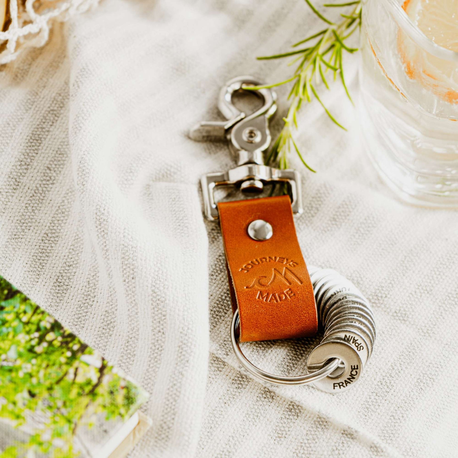 tan travel gift with leather travel keyring and engraved travel tokens on a white and cream striped blanket with green foliage around