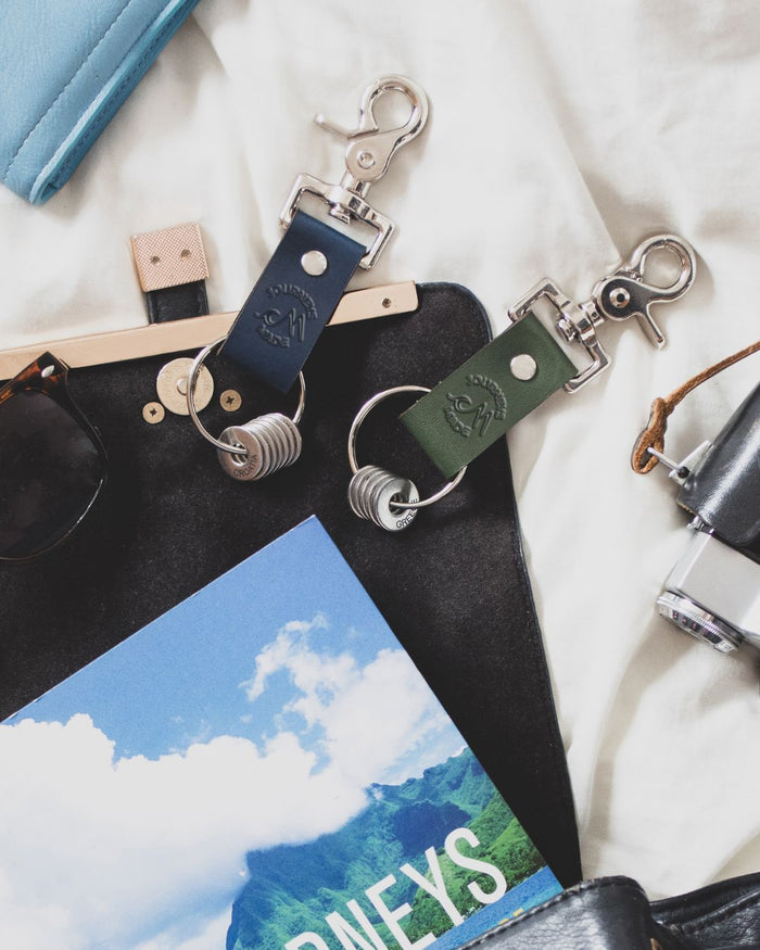 ocean blue and forest green travel keyring with engraved tokens laid on a black open handbag with a magazine titled Journeys