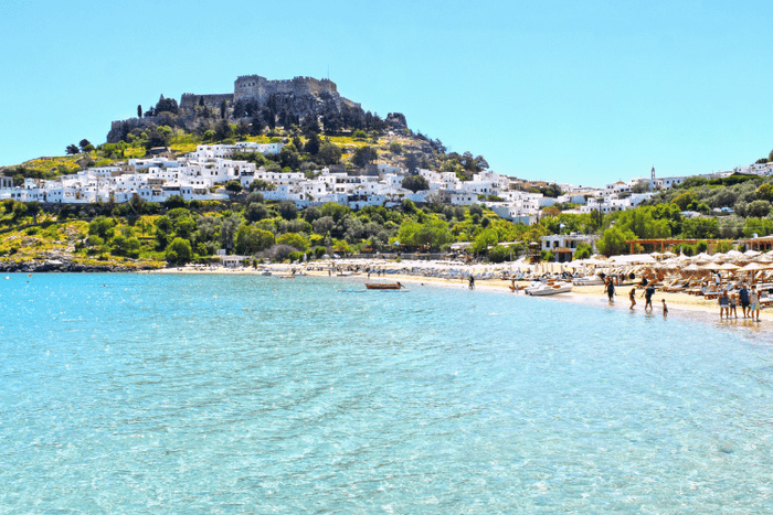 Stunning view of Lindos beach and ancient castle on Rhodes, Greece, featuring clear water and sunbathers.