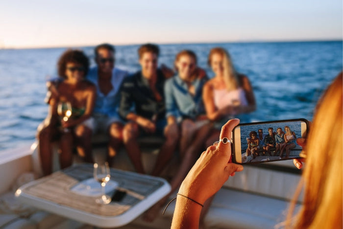 white woman holding a smartphone taking a photo of a group of friends on a yacht 