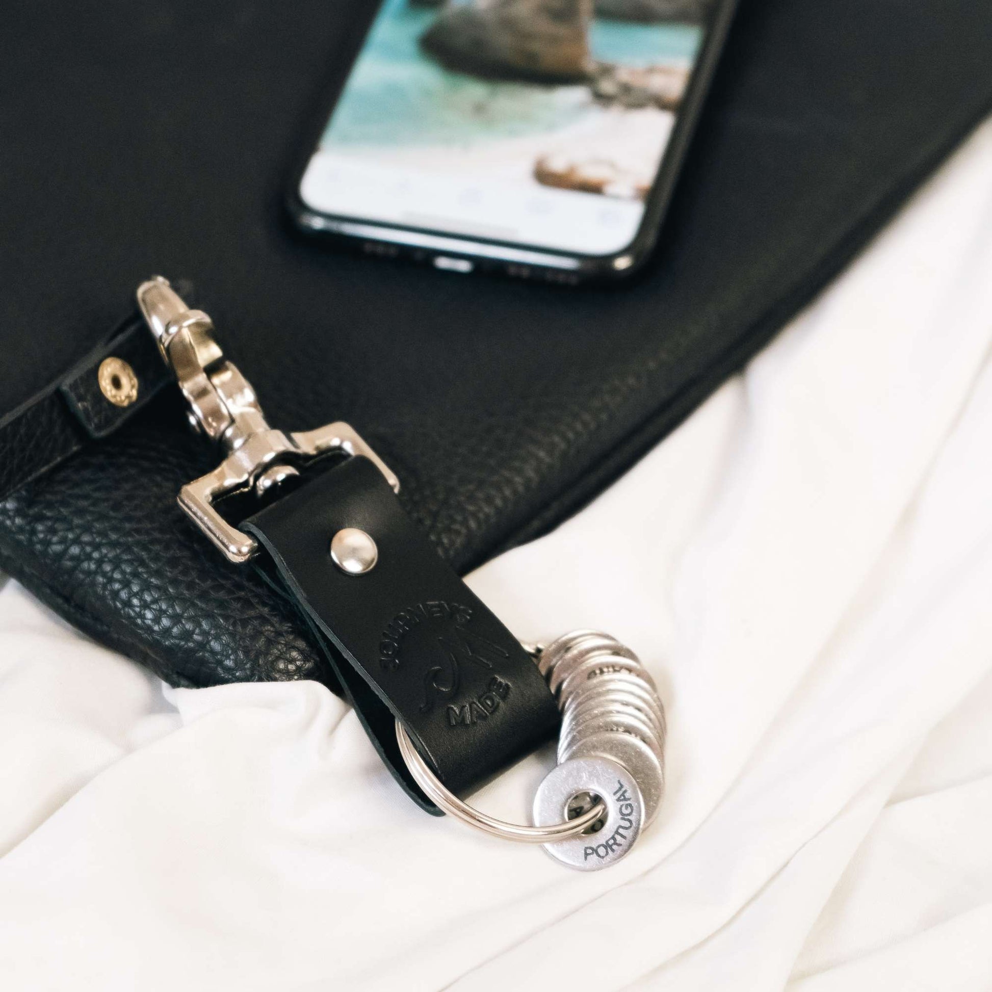 black travel gift with leather travel keyring and engraved travel tokens hooked onto a black bag with a photo showing a holiday photo in the background