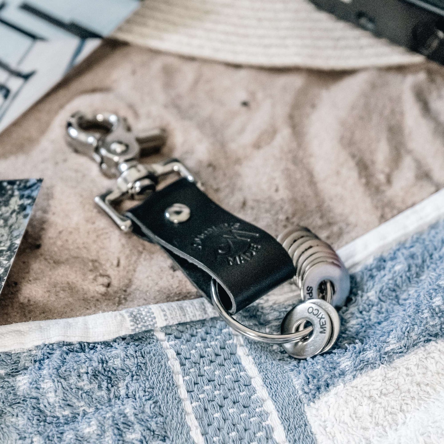 black travel gift with leather travel keyring and engraved travel tokens on a white and blue towel on sand with a sunhat and black camera in the background