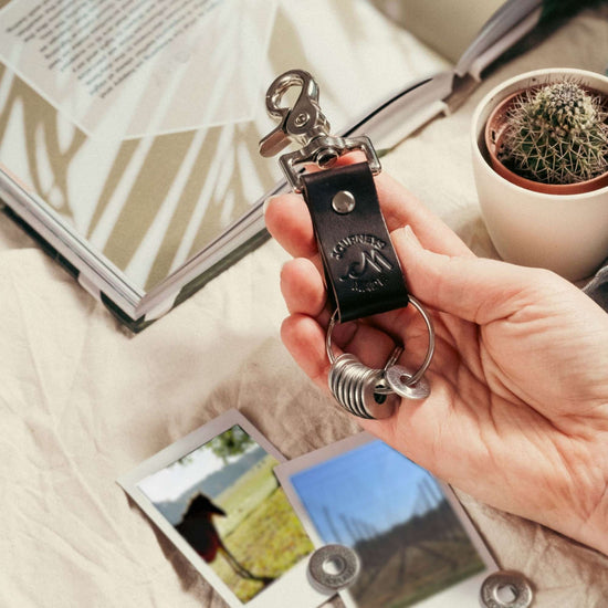 dark brown travel gift consisting of leather travel keyring and engraved travel tokens on the split ring with loose tokens on a table underneath, on the table are polaroid holiday photos