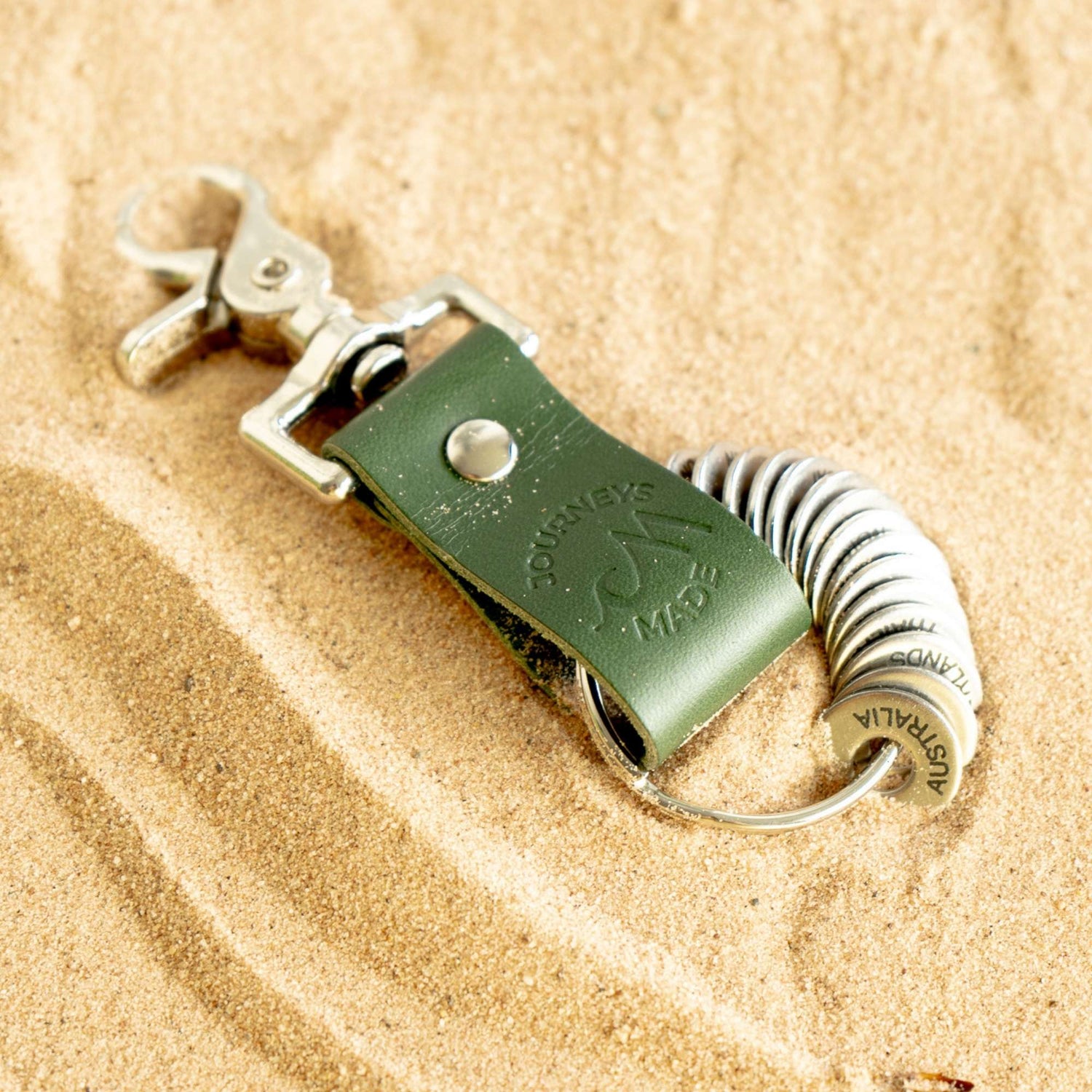 Forest green leather travel keychain with engraved destination tokens on sandy surface.