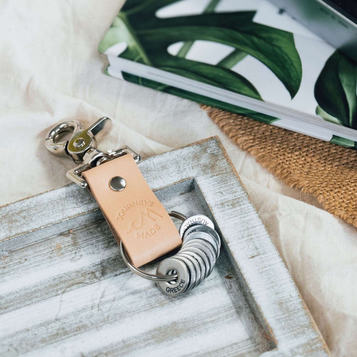 natural travel gift with leather travel keyring and engraved travel tokens on a white-washed wooden board with a leaf patterned book in the background