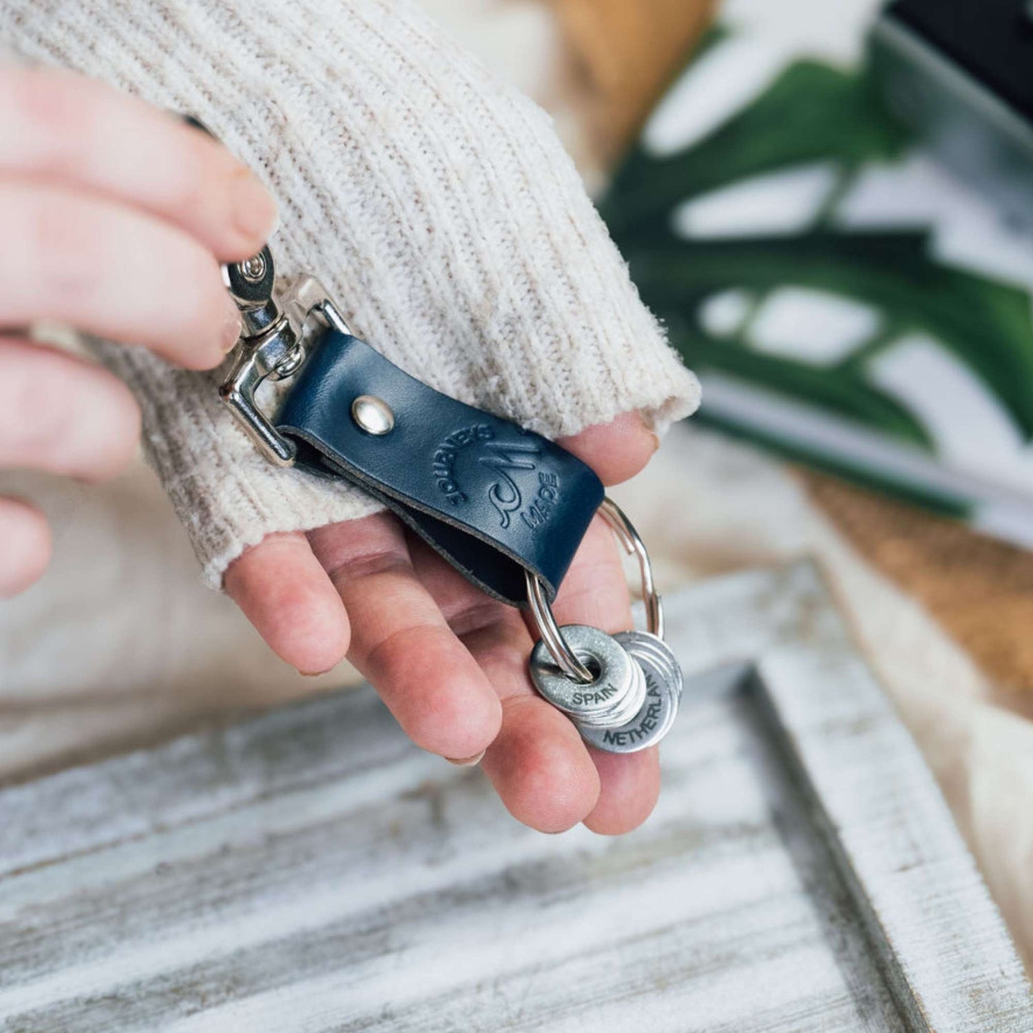 ocean blue travel gift consisting of leather travel keyring and engraved travel tokens on the split ring, being held in a white woman's hand wearing a white knitted jumper and a white wooden board in the background