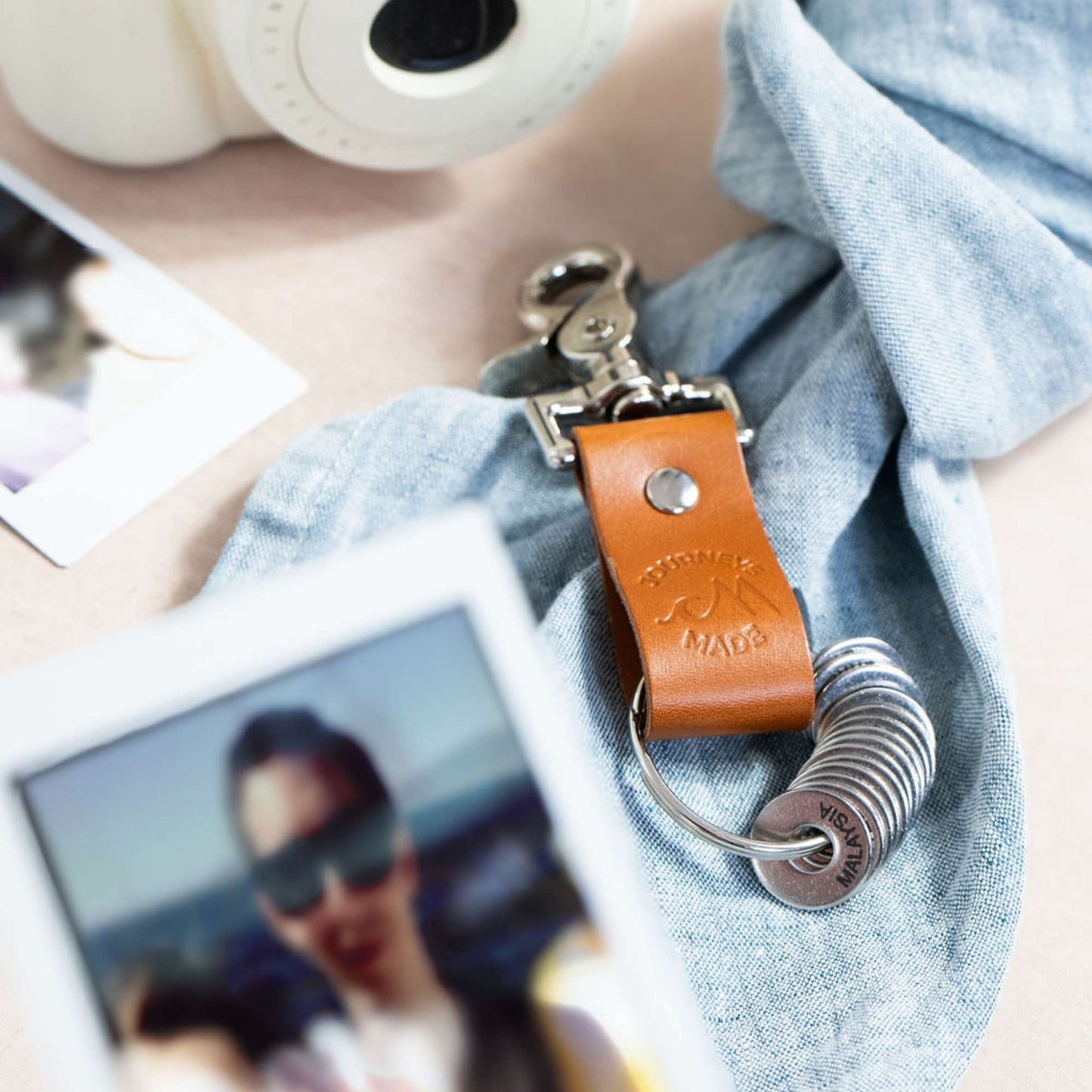 tan travel gift with leather travel keyring and engraved travel tokens on a blue denim tied cloth with a white polaroid camera and printed polaroid holiday photos